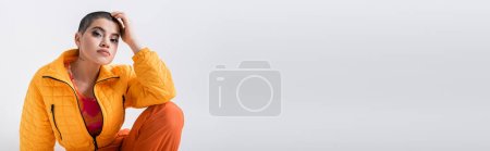 Photo for Urban fashion, outerwear, female model posing in casual attire, young woman with short hair and puffer jacket sitting on grey background, looking at camera, personal style, tattooed, banner - Royalty Free Image