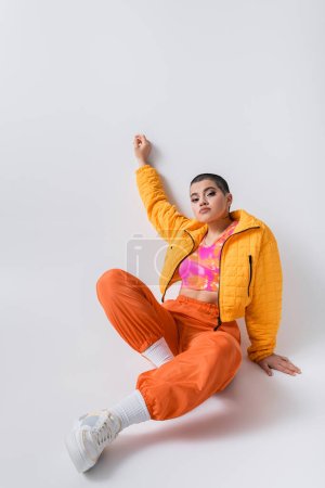 colorful clothes, outerwear, female model posing in casual attire, young woman with short hair and puffer jacket sitting on grey background, looking at camera, personal style, self expression 