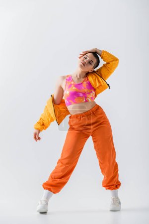 personal style, fashionable young woman with short hair posing in puffer jacket and orange pants on grey background, outerwear, modern subculture, youthful energy, full length 