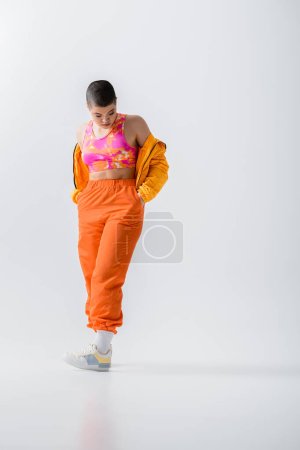 Moderne Subkultur, modische junge Frau mit kurzer Hose, die Hände in orangefarbenen Hosentaschen auf grauem Hintergrund, Pufferjacke. Oberbekleidung, jugendliche Energie, volle Länge 