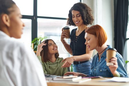 smiling multiracial woman holding paper cup with coffee to go and pointing with finger near multiethnic friends on blurred foreground in interest club, spending time in friendly diverse community