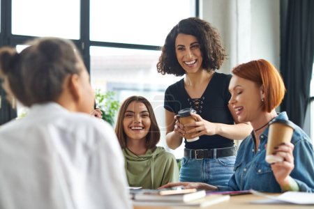 excited multiracial woman standing with paper cup of coffee to go during conversation with multiethnic female friends in interest club, spending time in friendly diverse community