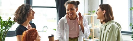 young and pleased african american woman showing new hoop earrings to happy girlfriends spending time in women interest club, gathering and leisure of diverse female team, banner