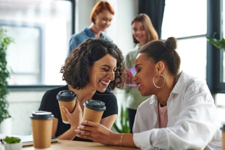 überglückliche afrikanisch-amerikanische und multirassische Freundinnen mit Coffee to go in der Hand und lachend mit geschlossenen Augen in der Nähe von Frauen auf verschwommenem Hintergrund im Interesse Club, Solidarität und Verständnis Konzept