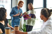 smiling woman pointing at magazine near amazed friend with mobile phone near multicultural women sitting with paper cups in interest club on blurred foreground, knowledge-sharing and communication Longsleeve T-shirt #664266578