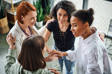 high angle view of joyous multiethnic girlfriends and smiling redhead psychologist joining hands as sign of unity during motivation session, moral support and mental wellness concept