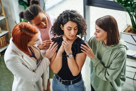 high angle view of psychologist and multiethnic female friends calming frustrated multiracial woman standing with closed eyes in consulting room, moral support and mental wellness concept