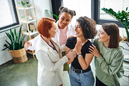 redhead motivation coach and smiling multicultural female friends embracing overjoyed multiracial woman during psychology session in consulting room, moral support and mental wellness concept