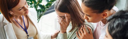 young depressed woman obscuring face and crying near motivation coach and multiethnic friends during psychology session in consulting room, problem-solving and mutual support concept, banner