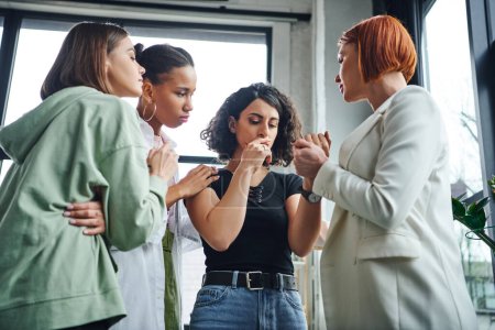 psychologist holding hand of frustrated multiracial woman and calming her together with multiethnic friends during motivation session in consulting room, problem-solving and mutual support concept