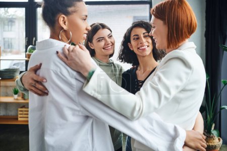 Photo for Happy and optimistic multicultural girlfriends embracing with redhead motivation coach and smiling at each other during supportive therapy in consulting room, female unity and support concept - Royalty Free Image