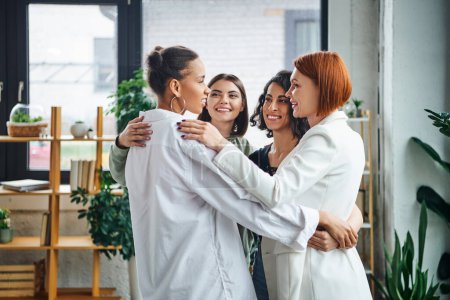 overjoyed multiethnic women and redhead motivation coach hugging and looking at each other while standing together in consulting room, female unity and support concept
