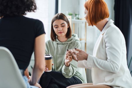 redhead psychologist holding hand of young and sad woman sitting with coffee to go during supportive therapy in consulting room, problem-solving and mental wellness concept