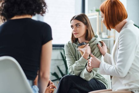 redhead psychologist calming young upset woman sitting in consulting room with takeaway drink near girlfriend on blurred foreground, understanding and mutual support concept
