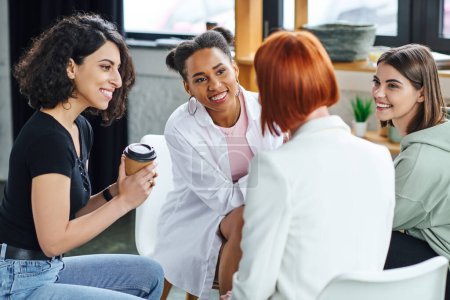multiracial woman sitting with coffee to go and listening to redhead psychologist together with optimistic multiethnic girlfriends during therapy, understanding, support and mental health concept