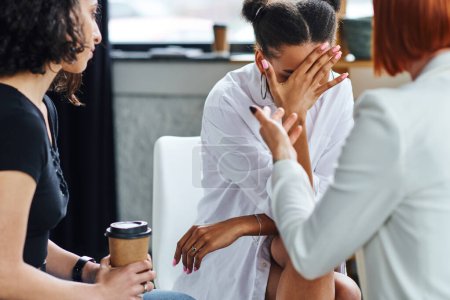 depressed african american woman obscuring face with hand and crying near psychologist and multiracial friend sitting with coffee to go in consulting room, problem-solving concept