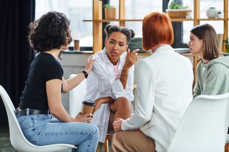 woman with coffee to go touching shoulder of african american girlfriend and supporting her during psychology session in consulting room, problem-solving and mental wellness concept