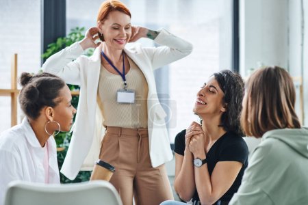 Photo for Pleased motivation coach smiling near diverse multicultural group and multiracial woman laughing with closed eyes during psychology session, understanding, support and mental health concept - Royalty Free Image