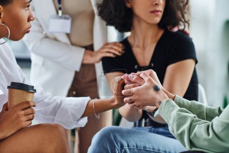 multiethnische Frauen mit Tätowierungen und Kaffee zu gehen Hand in Hand mit depressiver Freundin, während Beruhigung und Unterstützung während Psychologie-Sitzung, Problemlösung und mentales Wellness-Konzept