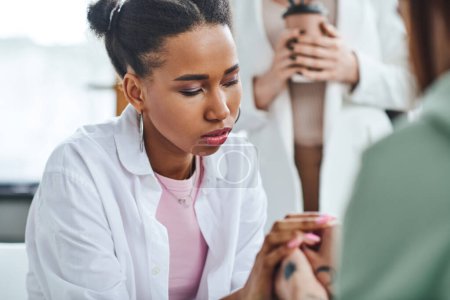 young and thoughtful african american woman holding hands of girlfriend during supportive therapy near psychologist standing with paper cup on blurred background, empathy and problem-solving concept