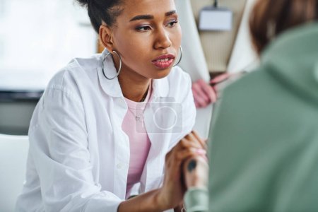 young and serious african american woman holding hands of friend near professional psychologist on blurred background in consulting room, empathy, solidarity and problem-solving concept