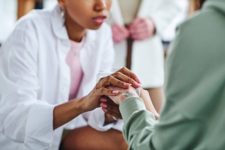 cropped view of african american woman holding hands of young friend during supportive therapy near psychologist on blurred background, empathy and problem-solving concept