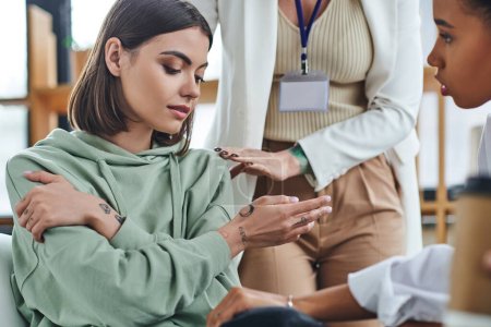 professional psychologist calming disappointed tattooed woman sitting near african american female friend on blurred foreground in consulting room, empathy and problem-solving concept