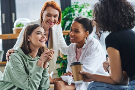 inspired and excited woman sitting and talking to multiethnic female friends and smiling psychologist during supportive session in consulting room, motivation and mental wellness concept
