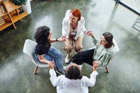 high angle view of joyful multiethnic female friends and redhead motivation coach sitting in circle and holding hands while talking on group therapy, communication and mental wellness concept