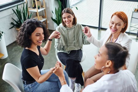 cheerful multicultural female friends and redhead motivation coach holding hands and communicating during group therapy in consulting room, friendship and mental wellness concept