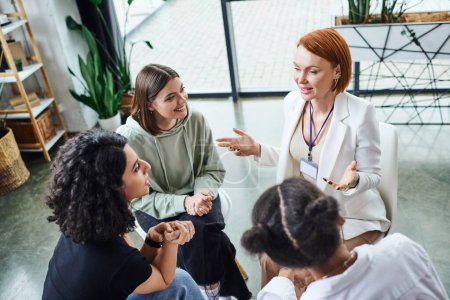 high angle view of redhead psychologist pointing with hands and talking to smiling multicultural women during motivation therapy in consulting room, friendship and mental wellness concept
