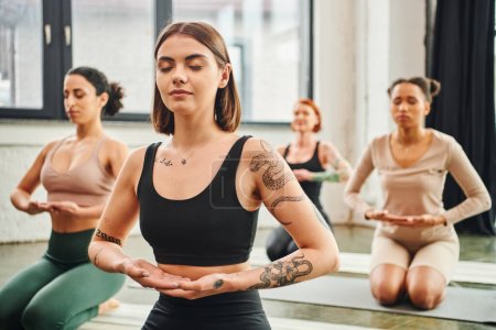 young and tattooed woman in sportswear meditating with closed eyes near diverse group of multiethnic friends on blurred background, inner peace and body awareness concept