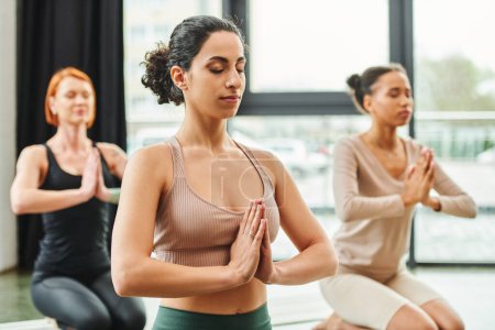 multiethnische Frau in Sportkleidung meditiert mit geschlossenen Augen und betenden Händen während eines Yoga-Kurses in der Nähe multiethnischer Freundinnen auf verschwommenem Hintergrund, innerem Frieden und Körperwahrnehmungskonzept