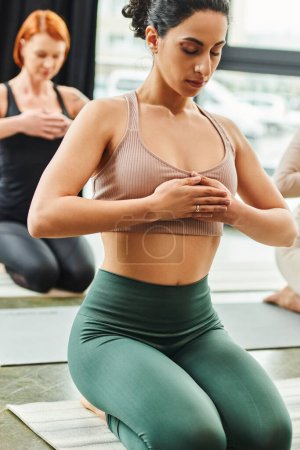 multiracial woman meditating with closed eyes and hands on chest while sitting in thunderbolt pose next to redhead girlfriend on blurred background, inner peace and body awareness concept