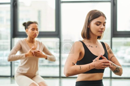 young and tattooed woman meditating with closed eyes and hands on chest during yoga class near african american girlfriend on blurred background, inner peace and body awareness concept