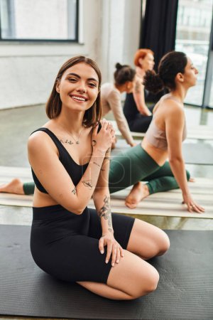 young and overjoyed tattooed woman in sportswear looking at camera while sitting near multicultural female friends meditating on yoga class, wellness and mental health concept