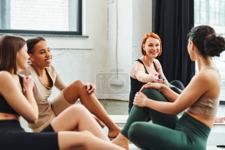 mujer feliz, pelirroja y tatuada señalando con el dedo y hablando con amigos multiculturales sentados en colchonetas de yoga en gimnasio, amistad, armonía y concepto de salud mental