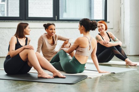diverse group of happy and smiling multiethnic female friends in sportswear sitting on yoga mats and talking in gym, friendship, harmony and mental health concept