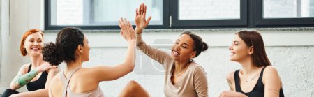 young, carefree and excited african american woman giving high five to friend near cheerful women during yoga class in gym, friendship, harmony and mental health concept, banner