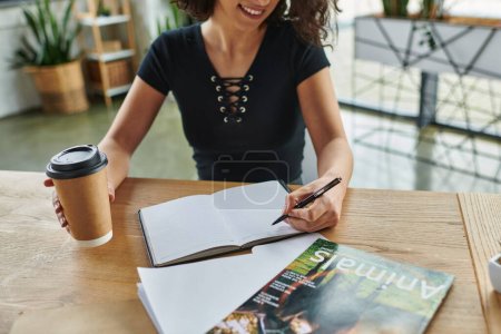 personal growth, self-education, cropped view of smiling multiracial motivation coach with coffee to go writing in notebook near magazine in women interest club, social activism concept