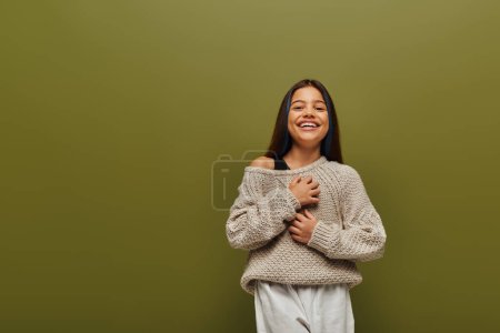 Chica preadolescente alegre en suéter de punto elegante y traje casual tocando el pecho mientras mira a la cámara y de pie aislado en verde, moda contemporánea para el concepto preadolescente 
