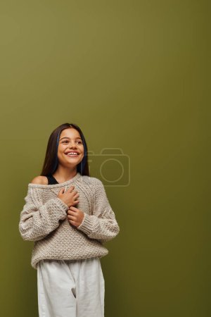 Chica preadolescente morena alegre en suéter de punto y traje casual tocando el pecho mientras mira hacia otro lado y de pie sobre fondo verde, moda contemporánea para el concepto preadolescente