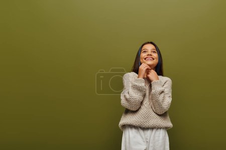 Chica preadolescente soñadora y sonriente con el pelo teñido en suéter de punto elegante mirando hacia otro lado mientras está de pie y posando aislado en verde, moda contemporánea para el concepto preadolescente