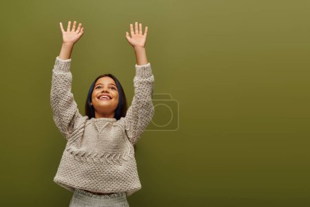 Morena positiva preadolescente niño con el pelo teñido en suéter de punto moderno levantando las manos y mirando hacia otro lado mientras está de pie aislado en verde, moda contemporánea para el concepto preadolescente