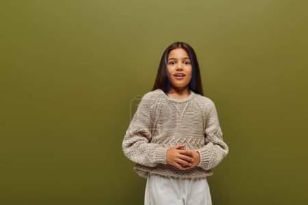 Photo for Excited brunette preteen girl with dyed hair in stylish knitted sweater and casual clothes looking at camera while standing isolated on green, contemporary fashion for preteen concept - Royalty Free Image