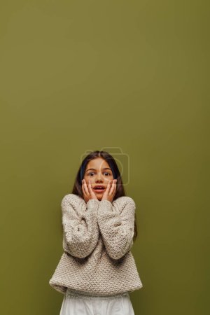 Sorprendido chica preadolescente morena con el pelo teñido en casual y elegante suéter de punto tocando las mejillas y mirando a la cámara aislada en verde, la moda contemporánea para el concepto preadolescente