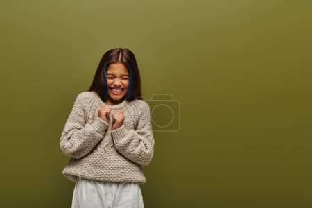 Muchacha preadolescente morena asustada en suéter de punto elegante y ropa casual cerrando los ojos mientras está de pie y posando aislado en verde, moda contemporánea para el concepto preadolescente