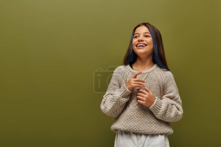 Happy and trendy brunette preteen girl with dyed strands of hair wearing modern knitted sweater and looking away while standing isolated on green, contemporary fashion for preteen concept