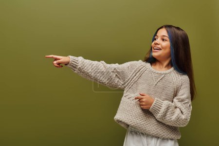 Chica preadolescente alegre y de moda con soportes teñidos de pelo con suéter de punto moderno y apuntando con el dedo aislado en verde, moda contemporánea para el concepto preadolescente