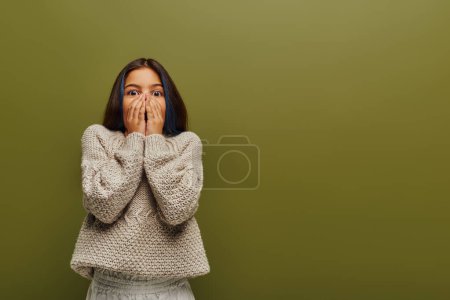 Scared preteen kid with dyed hair wearing stylish knitted and cozy sweater while covering face and looking at camera isolated on green, contemporary fashion for preteen concept